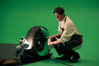 A man in a white shirt and apron crouches while using a power tool on a large tire in front of a green screen.