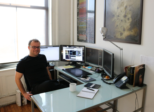 A man wearing glasses and a black t-shirt sits at a desk with three computer monitors, a telephone, and books.