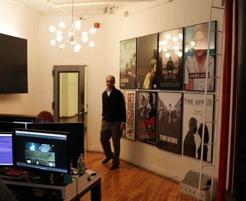 A man walks past a wall of framed TV show posters including The Wire and Nurse Jackie in an office.