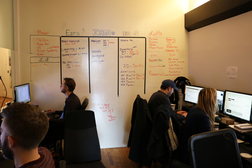 Office whiteboard with names and tasks, employees working at computers.