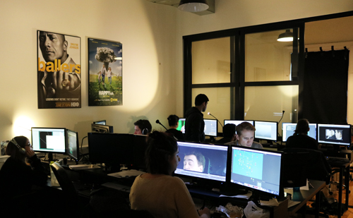 Several people are seated at desks, working on computers in a room with movie posters on the wall. One person is standing.