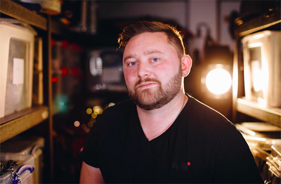 A man with a beard and short hair wearing a black t-shirt, looking at the camera in a dimly lit workshop.