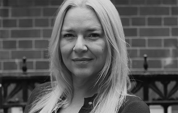 Black and white close-up portrait of a blonde woman smiling slightly in front of a brick wall.