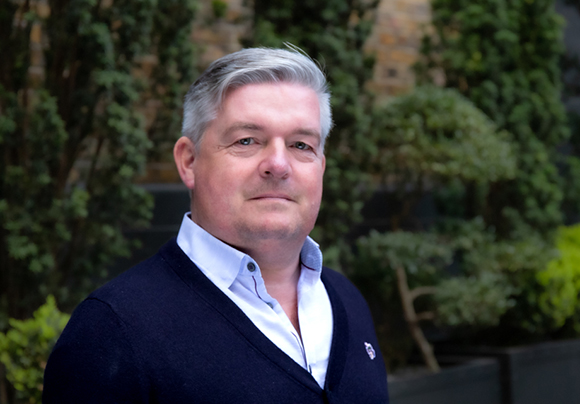 A man with grey hair wearing a navy cardigan over a light blue button-down shirt.