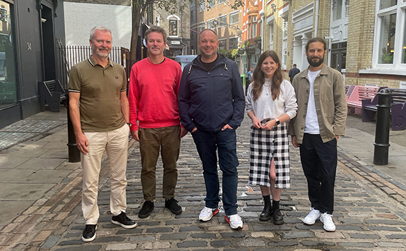Five people standing in a row on a cobblestone street in front of brick buildings.