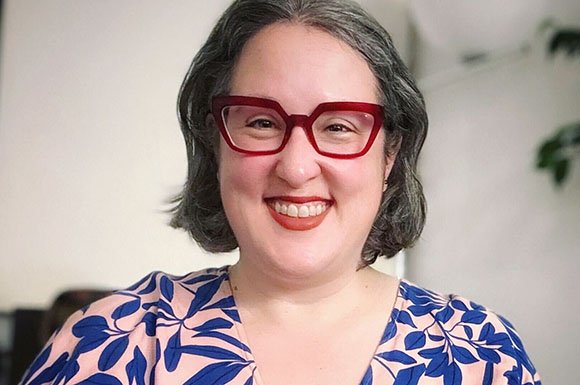 A smiling woman with short grey hair and thick red-rimmed glasses wearing a blue and pink patterned top.