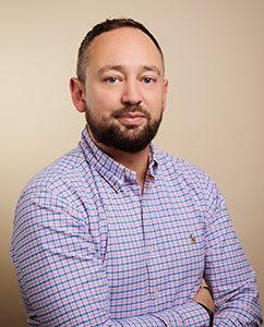 A man with a beard wearing a blue and pink checkered button-down shirt against a plain background.