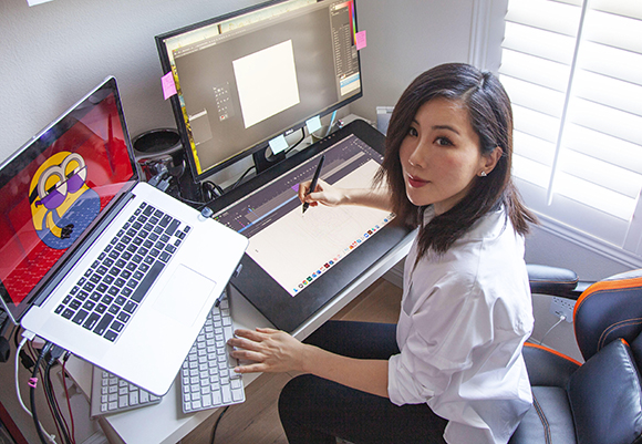 A woman sits at a desk using a stylus on a large tablet and a laptop with a colorful graphic.