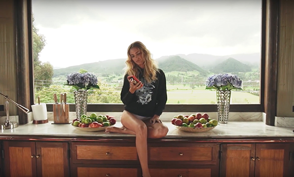 A woman with blonde hair sits on a marble kitchen counter, looking at her smartphone with a scenic view behind her.