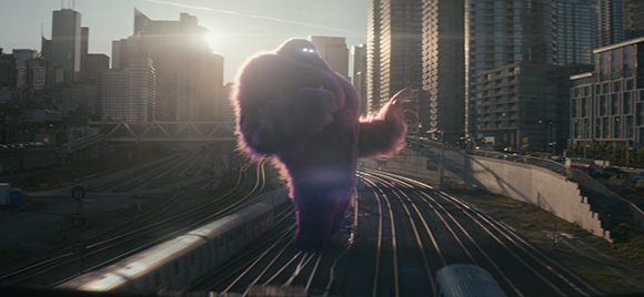 A large, glowing purple creature walks along train tracks through a sunlit urban cityscape.