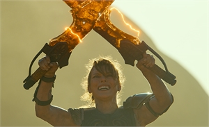 A smiling warrior holds two glowing, amber-colored axes high above her head against a bright sky.