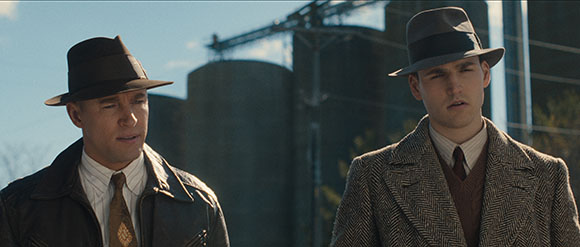 Two men in 1930s style fedoras and coats standing in front of large industrial silos.