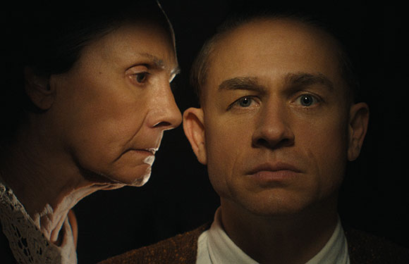 A woman in profile stares intensely at a man who looks directly forward against a dark background.