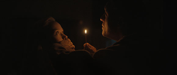 A person holds a lit candle between themselves and another person in a dark setting.