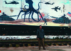 A person stands before a mural of a blue octopus-like creature, tanks, helicopters, and a jet.