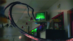 A long, dark, textured tentacle curves through a dimly lit room toward a green-glowing television screen.