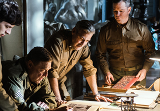 Four men in olive military uniforms lean over a table to study books and papers in a dimly lit room.