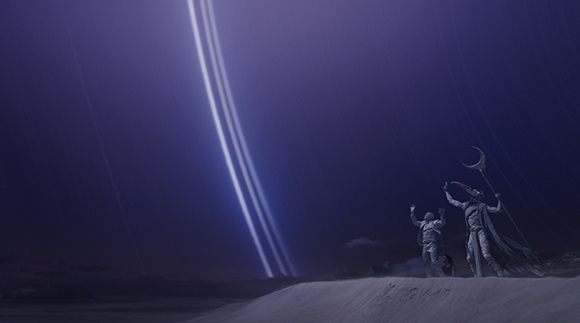 A person stands on a desert dune looking up at bright streaks of light in a dark purple sky.