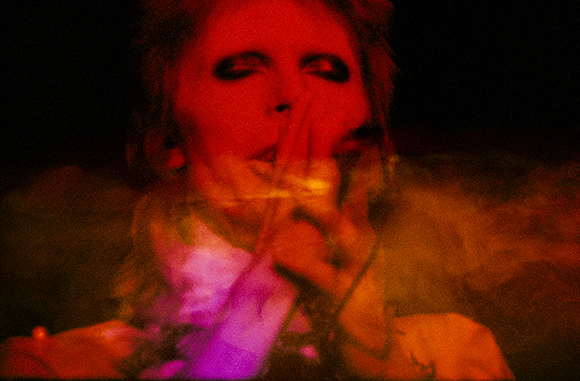 A person's face is bathed in intense red light amidst swirling smoke and shadows.
