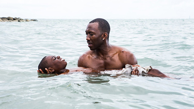 A shirtless man holds a person floating on their back in the ocean water.