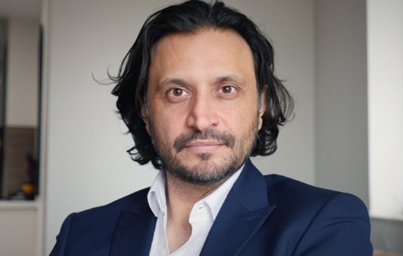 A man with dark wavy hair and a beard wearing a white shirt and navy blue blazer.