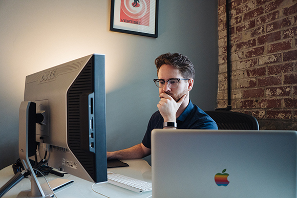 A man with glasses sits at a desk, looking thoughtfully at a large computer monitor.