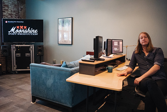 A person with long hair sits at a desk with dual monitors in a studio with a blue sofa and a large screen.