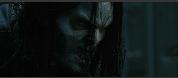 A close-up of a pale, menacing figure with dark hair, a beard, and a furrowed brow in dim lighting.