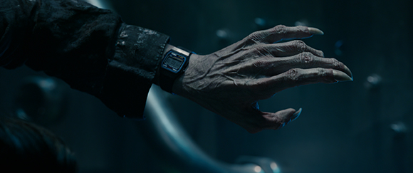 A pale, wrinkled hand with long, sharp fingernails wearing a black digital watch against a dark background.