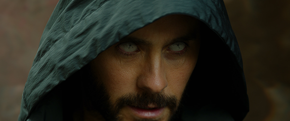 Close-up of a man with a dark beard and glowing white eyes peering out from under a dark hood.