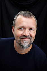 A man with short hair and a salt-and-pepper beard smiling against a dark background.