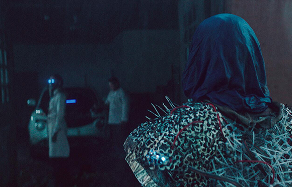 A person in a blue hood and leopard print clothing with spikes looks toward two blurry figures by a car.