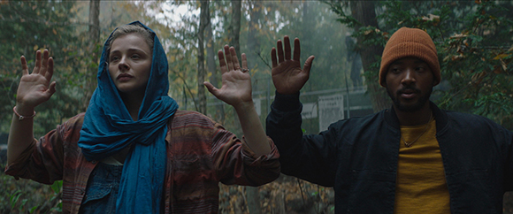 A woman in a blue headscarf and a man in an orange beanie raise their hands in a wooded area.