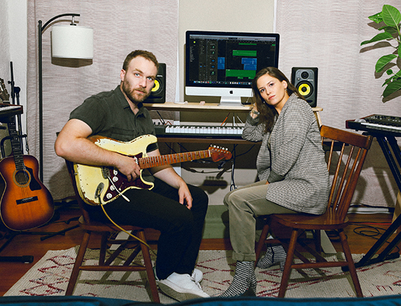 A man holding an electric guitar and a woman sitting in a music studio with recording equipment.
