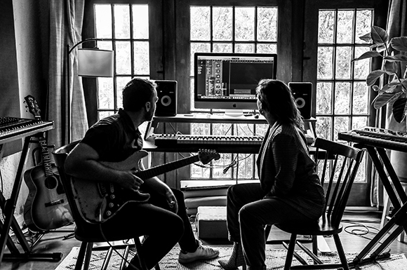 Two people sitting in a studio with a computer monitor, studio monitors, a keyboard, and guitars.