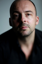 A close-up portrait of a bald man with light stubble looking directly at the camera.