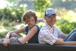 A woman with curly hair and a man in a baseball cap sit back-to-back on a bench in a sunny park.