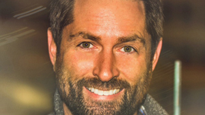 Close-up portrait of a smiling man with brown hair and a dark beard looking directly at the camera.