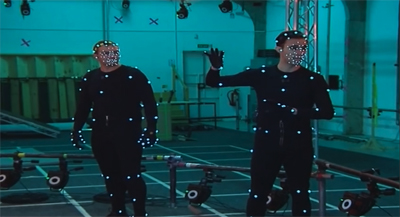 Two people wearing black motion capture suits with small reflective markers in a blue-lit studio.