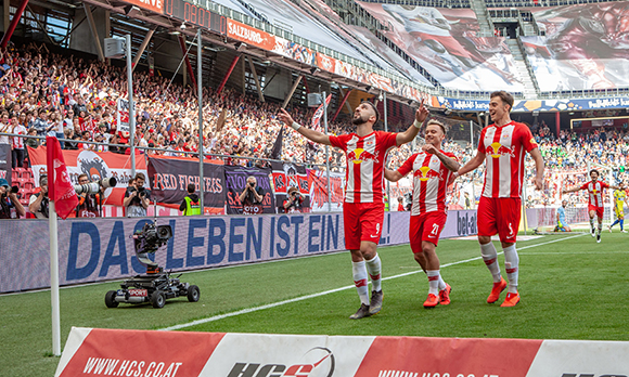 Three soccer players in red and white striped jerseys celebrate on a field before a large crowd.