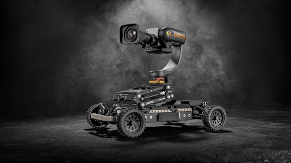 A black robotic rover with four rugged wheels carrying a mounted camera on a curved arm against a smoky background.