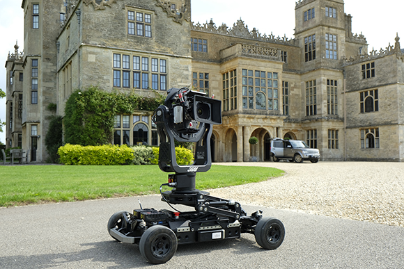 A black robotic camera stabilizer mounted on a motorized wheeled base in front of a large stone manor.