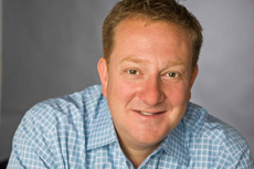 A smiling man with short light brown hair wearing a blue and white checkered button-down shirt.