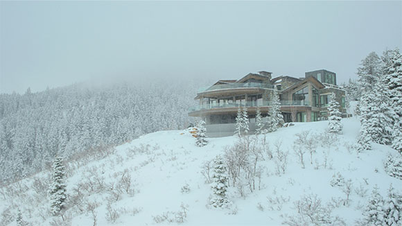 A large modern house with glass walls perched on a snow-covered mountain slope amidst evergreen trees.