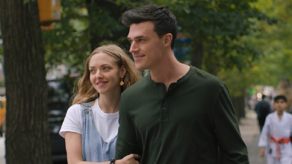 A smiling man in a dark green shirt and a woman in denim overalls walk along a tree-lined street.