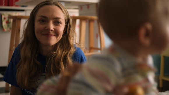 A woman with long blonde hair smiles warmly at a baby in the foreground.