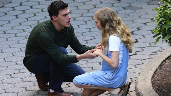 A man in a green shirt holds the hands of a woman in a blue denim dress while kneeling on a stone walkway.