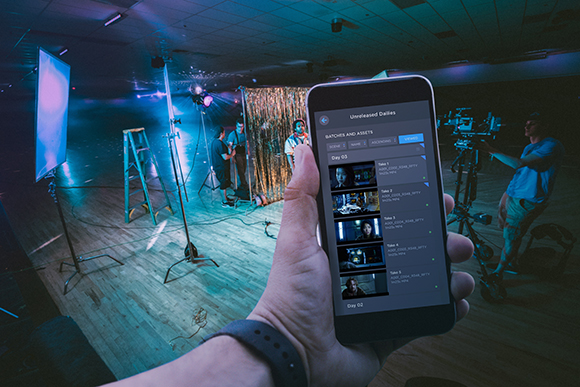 Hand holding a smartphone showing a video list in front of a film set with lights and crew.