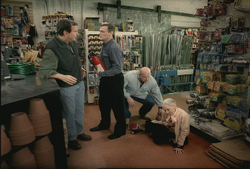 Four men stand and crouch in a hardware store aisle surrounded by tools and supplies.
