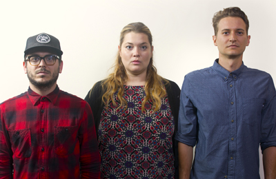 A man in a red plaid shirt, a woman in a patterned top, and a man in a blue button-down shirt stand together.